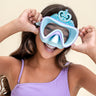 girl smiling and grabbing sides of turquoise crown kids' swim mask. swim mask showing silicone princess crown above purple frame and a light blue gasket 