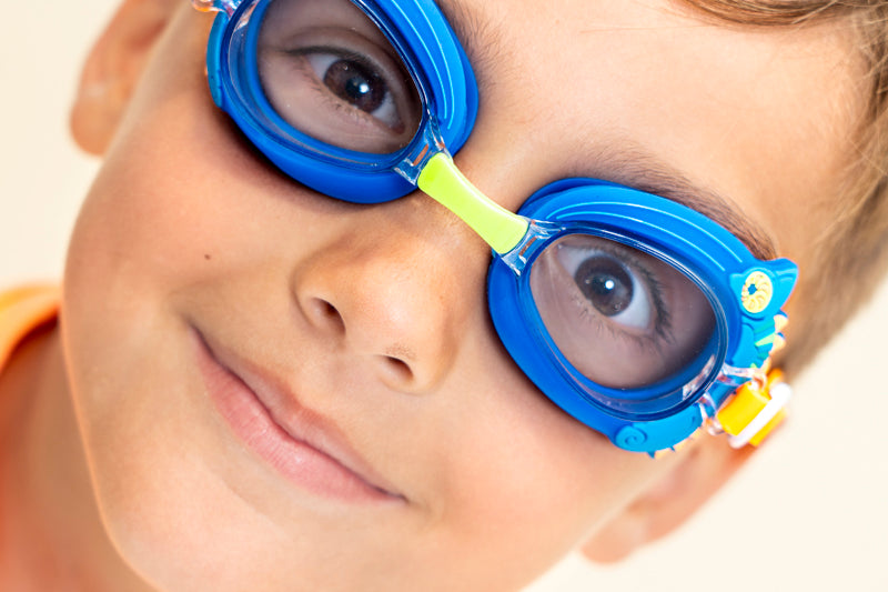 boy smiling wearing mini chameleon kids swim goggles featuring a blue frame with silicone chameleon decorations wrapping the frame and a lime green nose piece