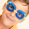boy smiling wearing mini chameleon kids swim goggles featuring a blue frame with silicone chameleon decorations wrapping the frame and a lime green nose piece