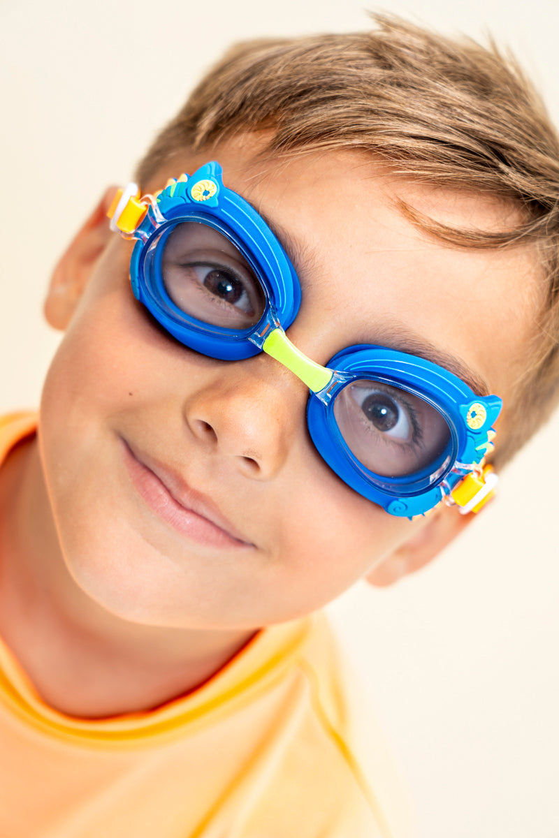 boy smiling wearing mini chameleon kids swim goggles featuring a blue frame with silicone chameleon decorations wrapping the frame and a lime green nose piece
