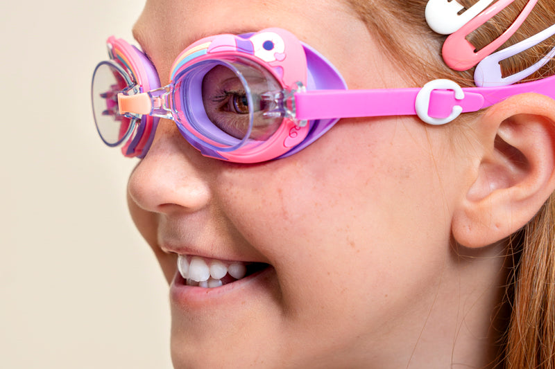 Angled shot of girl smiling wearing mini flamingo kids' swim goggles featuring a purple and pink colorway with a flamingo decoration wrapping around the frame with a peach colored nose piece 