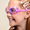 Angled shot of girl smiling wearing mini flamingo kids' swim goggles featuring a purple and pink colorway with a flamingo decoration wrapping around the frame with a peach colored nose piece 