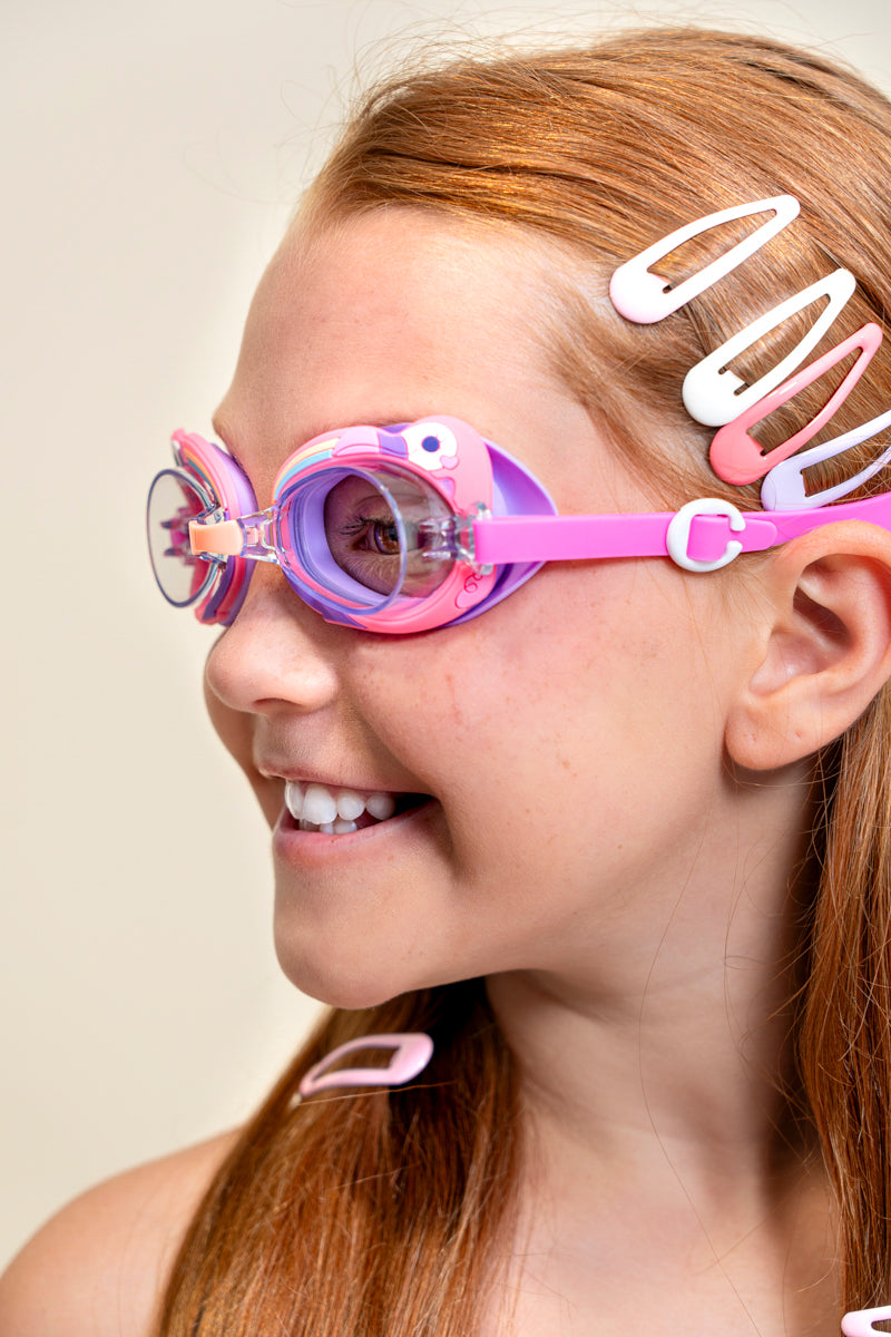 Angled shot of girl smiling wearing mini flamingo kids' swim goggles featuring a purple and pink colorway with a flamingo decoration wrapping around the frame with a peach colored nose piece 