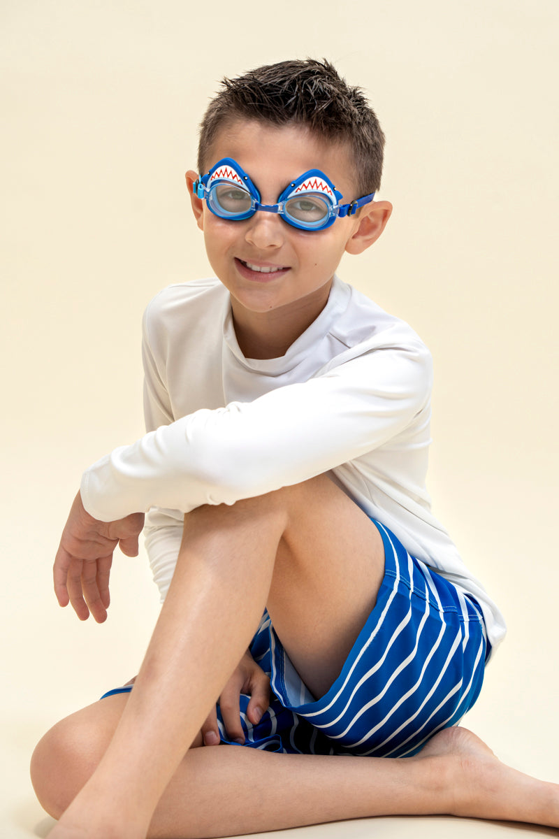 boy smiling and posing wearing mini mako shark kids swim goggles featuring a blue swim goggle with a silicone shark wrapping around the top of the lenses with a blue nose piece and blue strap