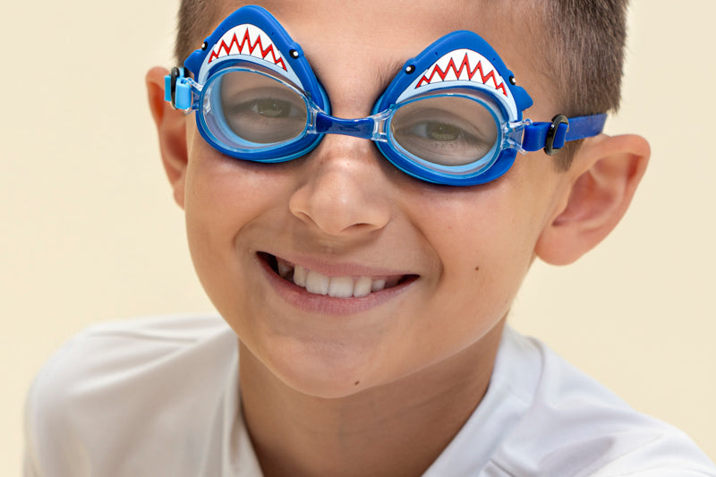 boy smiling wearing mini mako shark kids swim goggles featuring a blue swim goggle with a silicone shark wrapping around the top of the lenses with a blue nose piece and blue strap