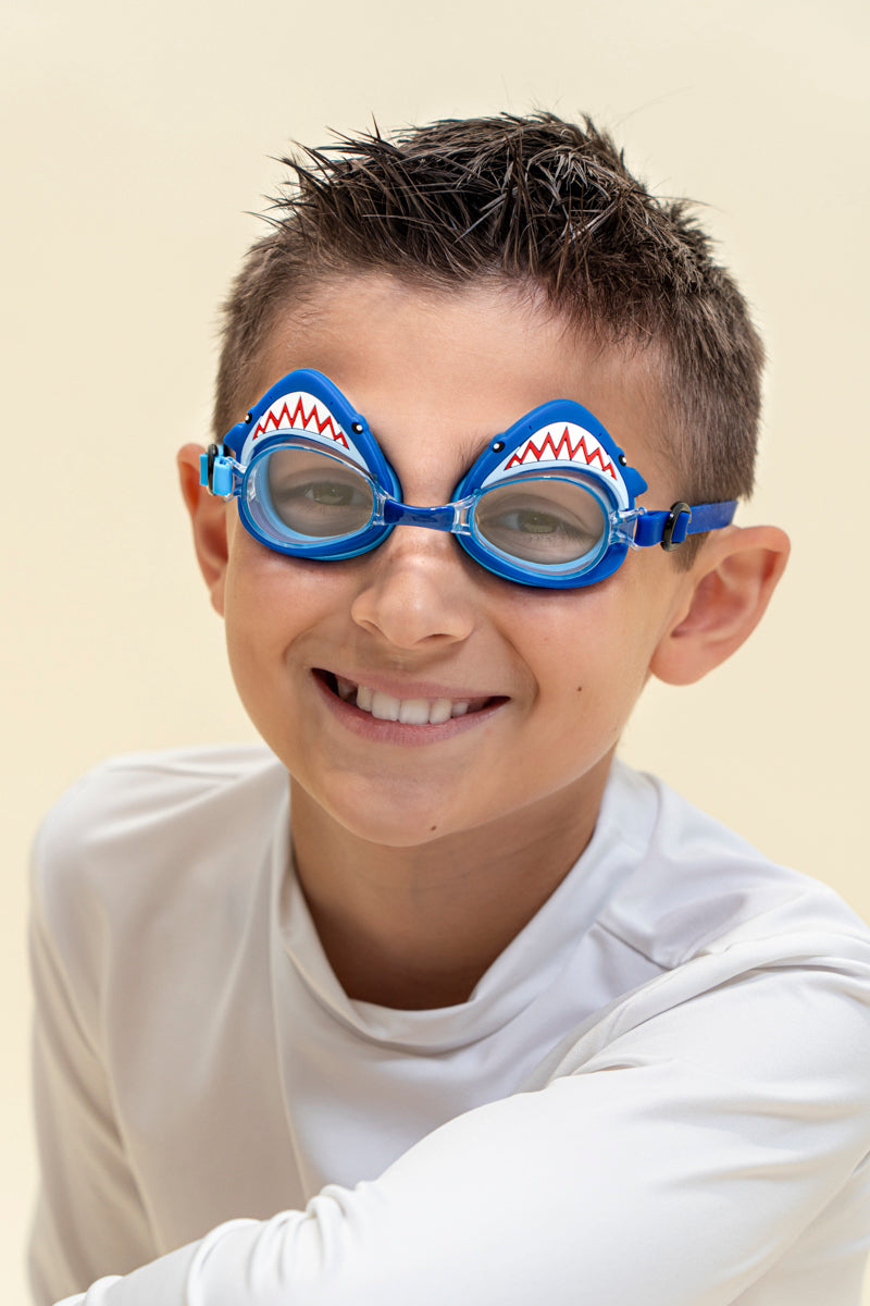 boy smiling wearing mini mako shark kids swim goggles featuring a blue swim goggle with a silicone shark wrapping around the top of the lenses with a blue nose piece and blue strap