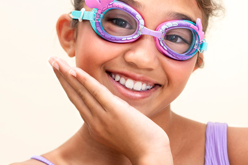girl smiling and posing wearing mini mermaid kids swim goggles which feature a mermaid tail inspired texture around the frame with a silicone mermaid tail coming off the side of the lens and a purple/turquoise colorway