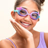 girl smiling and posing wearing mini mermaid kids swim goggles which feature a mermaid tail inspired texture around the frame with a silicone mermaid tail coming off the side of the lens and a purple/turquoise colorway