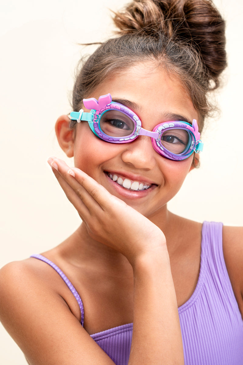 girl smiling and posing wearing mini mermaid kids swim goggles which feature a mermaid tail inspired texture around the frame with a silicone mermaid tail coming off the side of the lens and a purple/turquoise colorway