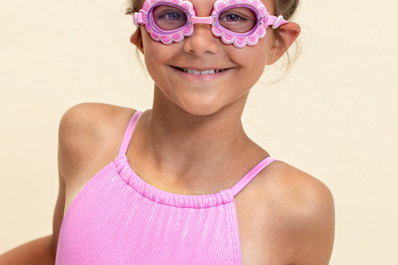 girl smiling wearing mini pink petals kids swim goggles featuring a pink swim goggle with pink petals wrapped around the lenses with a pink strap and nose piece