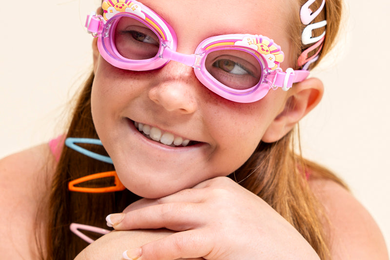girl sitting and smiling wearing mini pink sunshine kids swim goggles which feature a pink swim goggle with a silicone sunshine, clouds, and rainbow silicone decoration above the lens