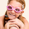 girl sitting and smiling wearing mini pink sunshine kids swim goggles which feature a pink swim goggle with a silicone sunshine, clouds, and rainbow silicone decoration above the lens