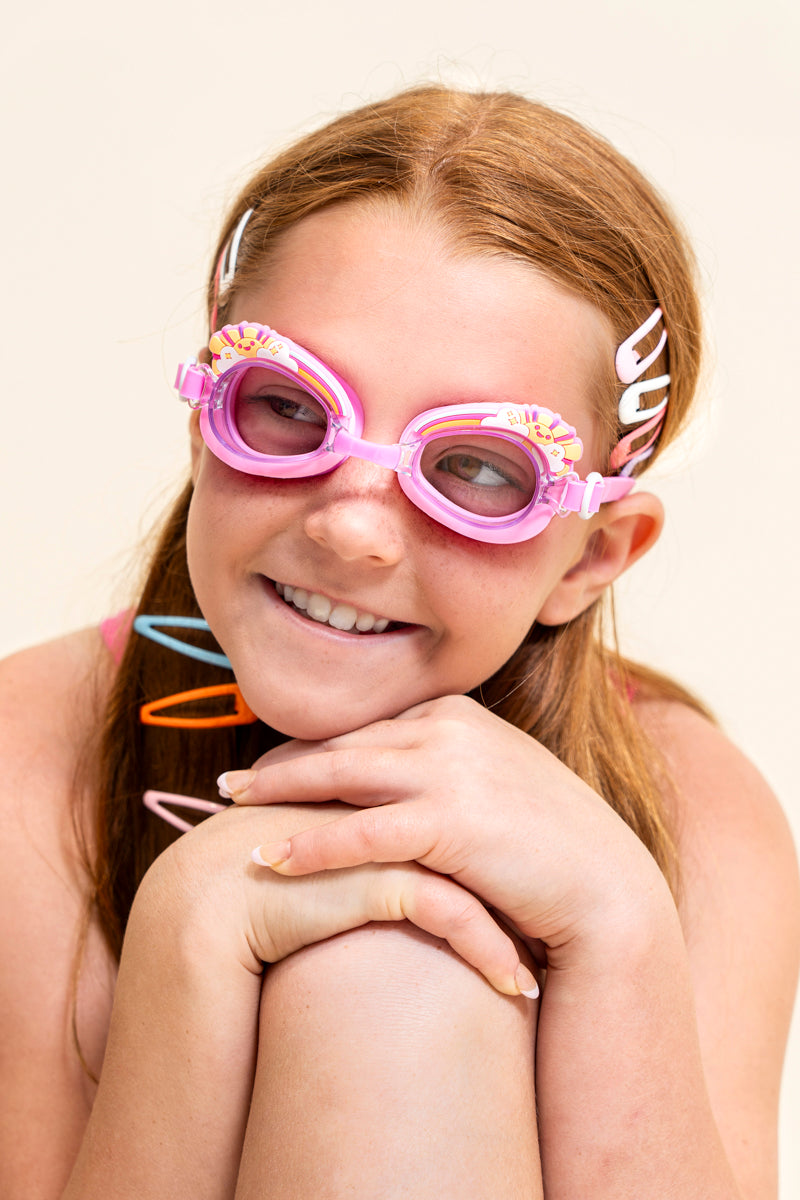 girl sitting and smiling wearing mini pink sunshine kids swim goggles which feature a pink swim goggle with a silicone sunshine, clouds, and rainbow silicone decoration above the lens