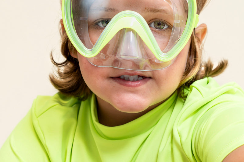 boy sitting smiling wearing splash in color green kids' swim mask showing a clear gasket and nose covering featuring a lime green frame and strap