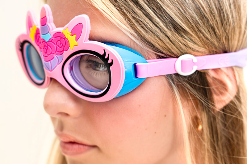 angled photo of girl wearing flex pink unicorn kids swim goggles featuring an all silicone flexible pink frame with a unicorn horn and ears with a flower crown on the silicone. Also featuring a blue gasket and pink strap