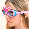 angled photo of girl wearing flex pink unicorn kids swim goggles featuring an all silicone flexible pink frame with a unicorn horn and ears with a flower crown on the silicone. Also featuring a blue gasket and pink strap