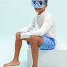 boy sitting and smiling wearing shark bite kids' swim mask showing shark teeth screen printed on the lens and a silicone shark about the lens also showing a light blue gasket and nose piece