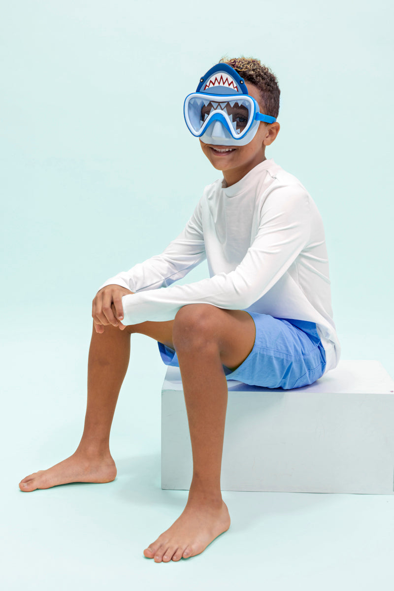 boy sitting and smiling wearing shark bite kids' swim mask showing shark teeth screen printed on the lens and a silicone shark about the lens also showing a light blue gasket and nose piece