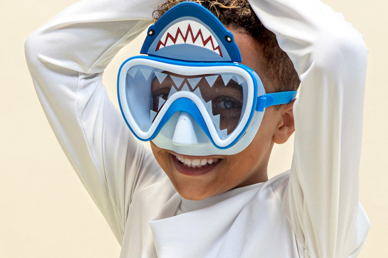 boy smiling posing with hands above head like a shark fin pose wearing shark bite kids' swim mask showing shark teeth screen printed on the lens and a silicone shark about the lens also showing a light blue gasket and nose piece