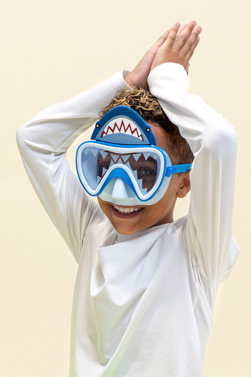 boy smiling posing with hands above head like a shark fin pose wearing shark bite kids' swim mask showing shark teeth screen printed on the lens and a silicone shark about the lens also showing a light blue gasket and nose piece