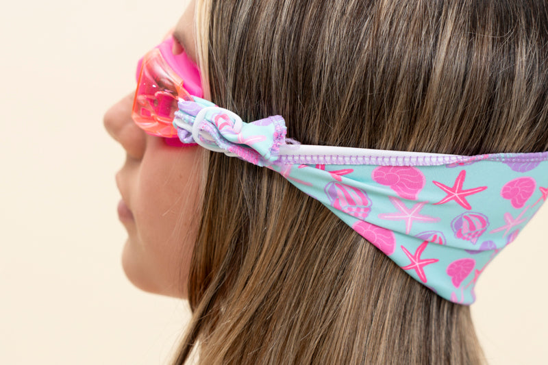 girl modeling soft strap coastal pink kids swim goggles which feature a turquoise colored soft strap with pink and purple coastal decorations such as seashells and starfish this shot features the soft strap fanned out across the back of the head