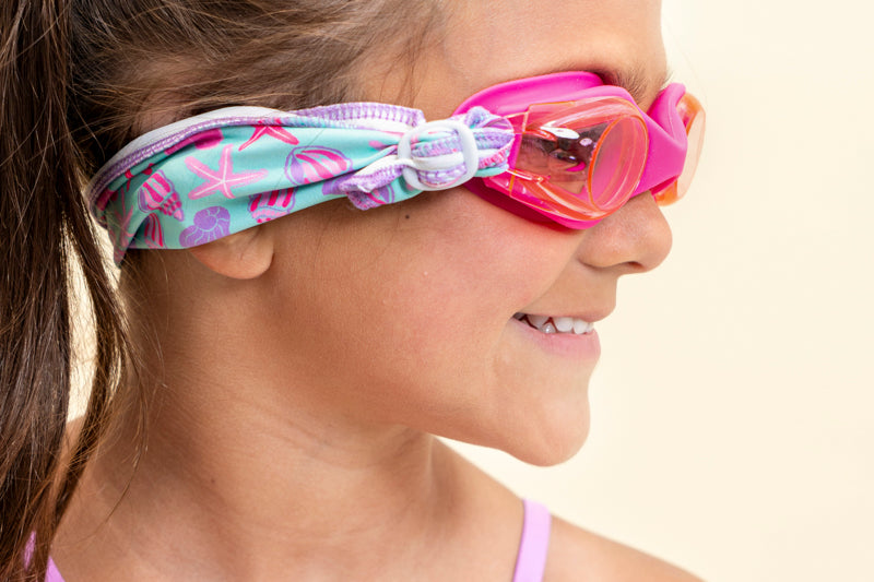 girl modeling soft strap coastal pink kids swim goggles which feature a bright pink swim goggle and a turquoise colored soft strap with pink and purple coastal decorations such as seashells and starfish this shot features the soft strap scrunched up across the back of the head