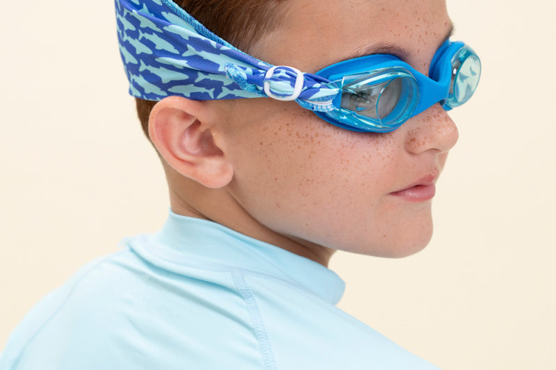 boy wearing soft strap shark blue kids swim goggles which feature a soft strap fanned out across the back of his head. the swim goggles feature a blue swim goggle and royal blue soft strap with light blue shark silhouettes  