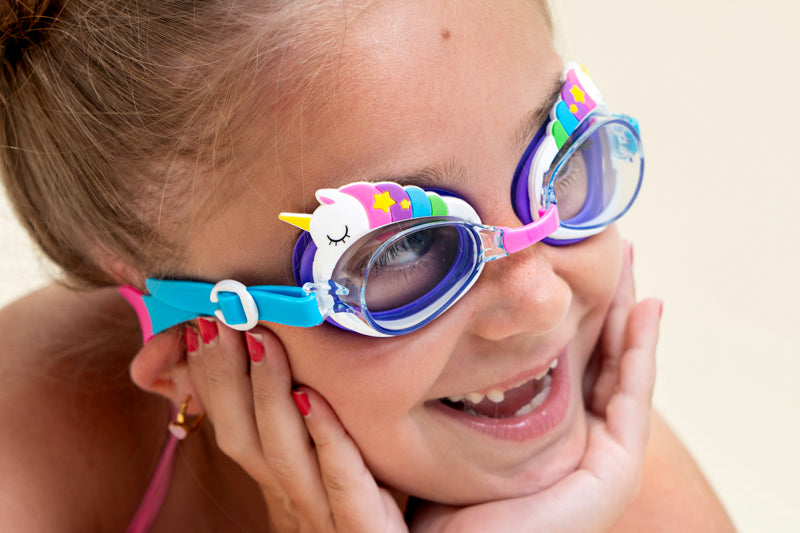 girl posing and smiling wearing mini unicorn kids swim goggles which feature  a silicone unicorn decoration wrapping around the top of the frame on a purple gasket and blue and pink strap with a pink nose piece