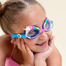 girl posing and smiling wearing mini unicorn kids swim goggles which feature  a silicone unicorn decoration wrapping around the top of the frame on a purple gasket and blue and pink strap with a pink nose piece
