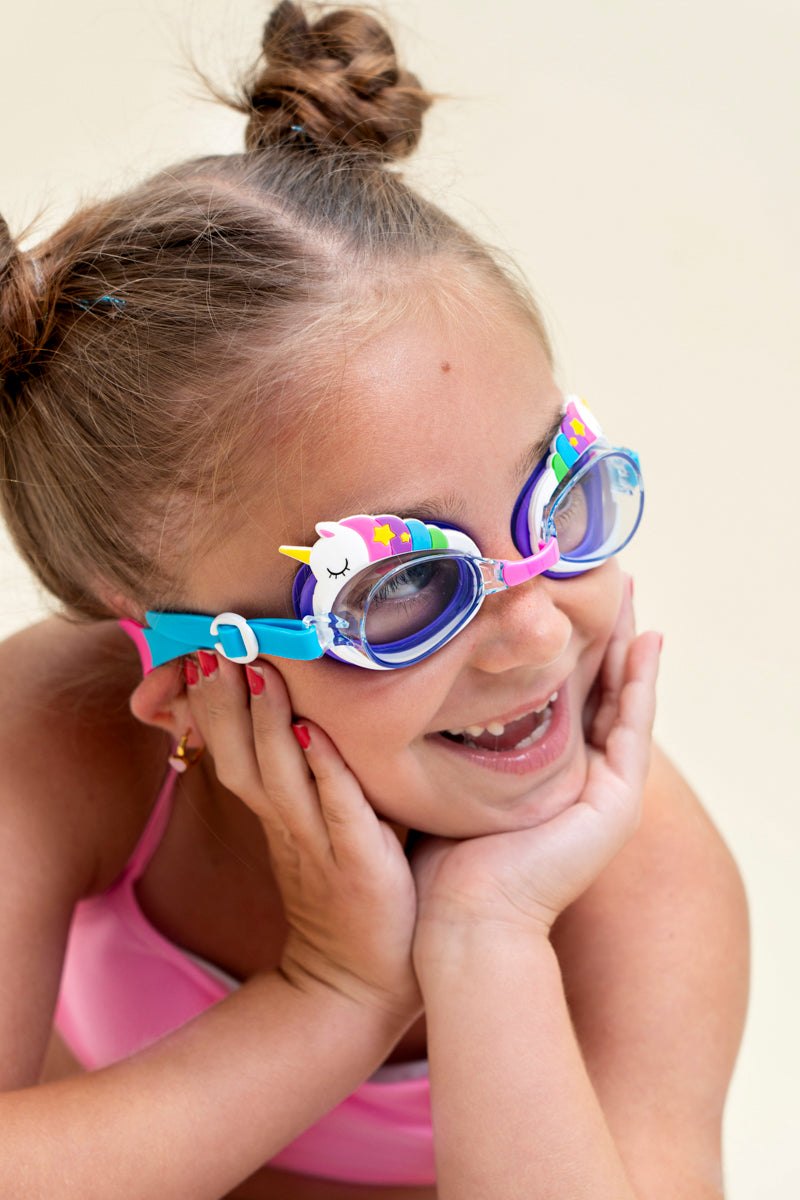 girl posing and smiling wearing mini unicorn kids swim goggles which feature  a silicone unicorn decoration wrapping around the top of the frame on a purple gasket and blue and pink strap with a pink nose piece