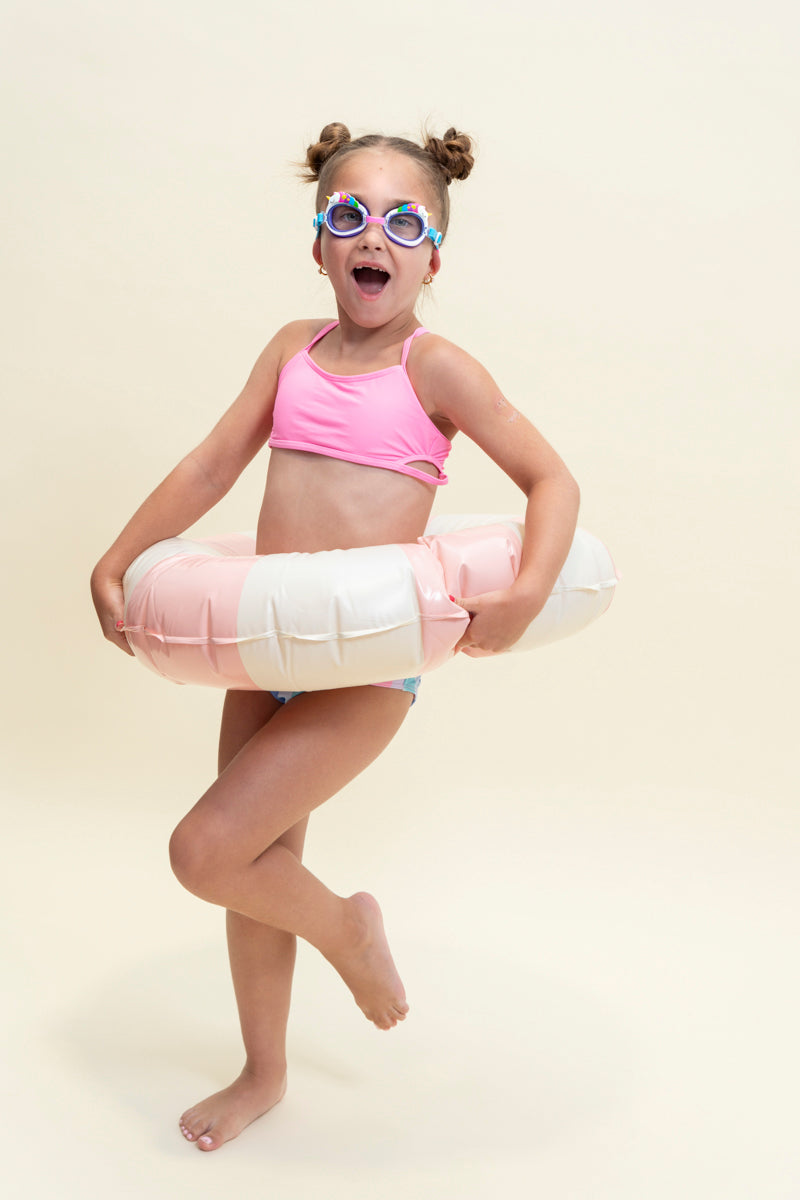 girl posing in an inner tube, smiling wearing mini unicorn kids swim goggles which feature  a silicone unicorn decoration wrapping around the top of the frame on a purple gasket and blue and pink strap with a pink nose piece