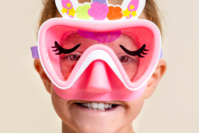 girl smiling wearing pink unicorn kids swim mask featuring a silicone unicorn horn and ears over the frame, a unicorn wink ;) print on the lens and a light pink gasket and nose covering