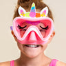 girl smiling wearing pink unicorn kids swim mask featuring a silicone unicorn horn and ears over the frame, a unicorn wink ;) print on the lens and a light pink gasket and nose covering