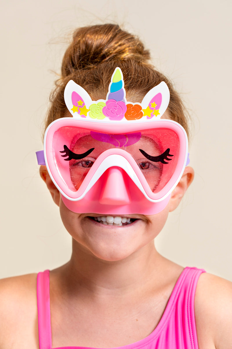 girl smiling wearing pink unicorn kids swim mask featuring a silicone unicorn horn and ears over the frame, a unicorn wink ;) print on the lens and a light pink gasket and nose covering