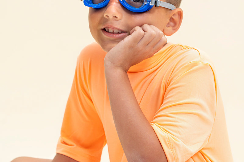 boy sitting and smiling wearing mini race car kids swim goggles which feature a blue swim goggle with silicone race car decorations across the top of both lens with a grey strap and red nose piece