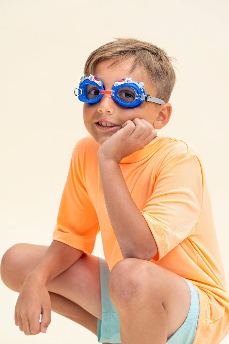 boy sitting and smiling wearing mini race car kids swim goggles which feature a blue swim goggle with silicone race car decorations across the top of both lens with a grey strap and red nose piece